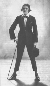 Berber standing astride wearing a tuxedo and carrying a cane. She has dramatic make-up and wears a monocle in one eye.
